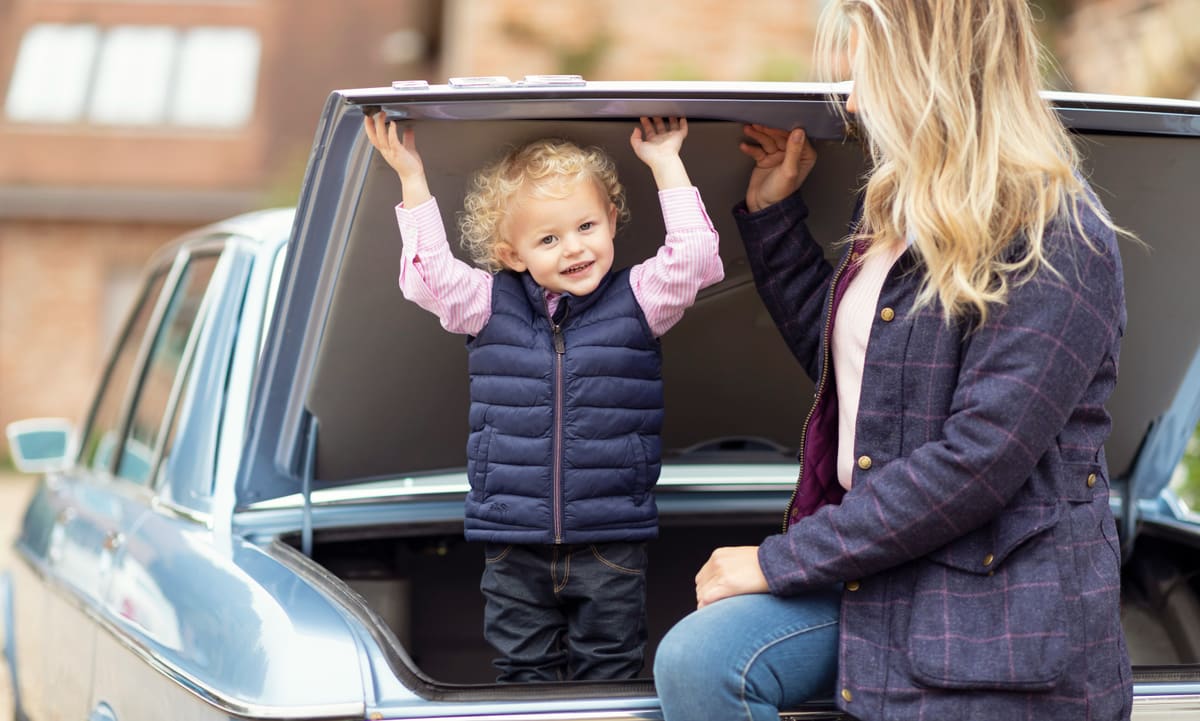 A smiling toddler with his arms up in the open boot of the family car, his mum sitting beside him